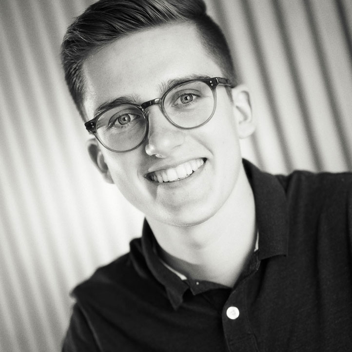 Junger Mann mit Brille und Hemd lächelt in die Kamera, schwarz-weiß Porträt vor unscharfem Hintergrund.
