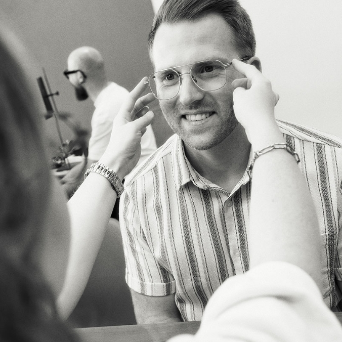 Person mit Brille lächelt, während jemand die Brille anpasst. Im Hintergrund sitzt eine andere Person. Schwarz-weiß.