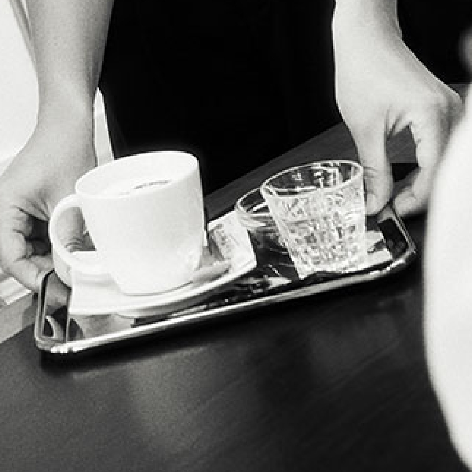 Person hält Tablett mit Kaffeetasse und Wasserglas auf dunklem Tisch. Schwarz-Weiß-Fotografie.