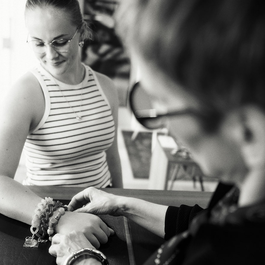 Zwei Frauen, von denen eine Armbänder an der Handgelenk der anderen begutachtet, in einem hellen Raum. Schwarz-Weiß-Foto.