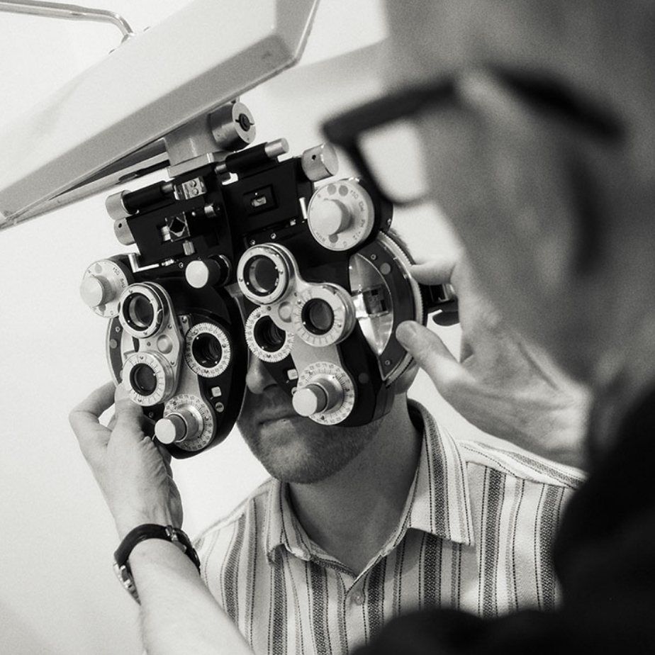 Person bei Sehtest, Optiker justiert Phoropter. Monochromes Bild mit Fokus auf Augenuntersuchungsgerät.