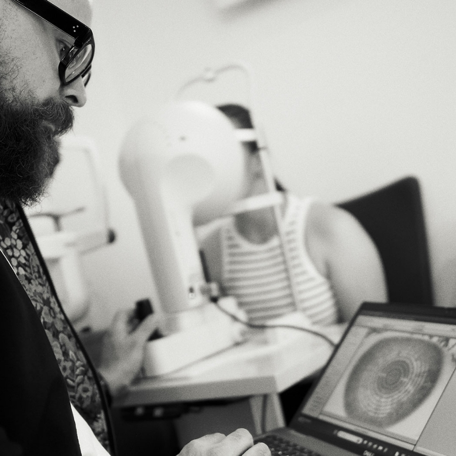 Optiker analysiert mit Brille ein Augenuntersuchungsergebnis auf einem Laptop, während Patientin im Hintergrund sitzt.