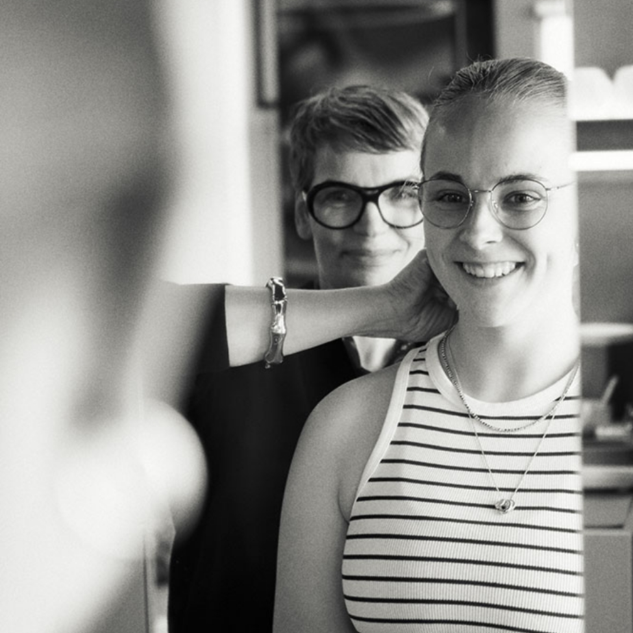 Zwei Frauen mit Brillen lächeln in einem Spiegel. Die vordere Frau trägt ein gestreiftes Top und eine Halskette. Schwarz-Weiß-Foto.