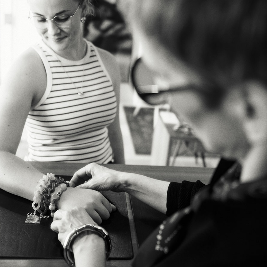 Zwei Personen, eine blickt auf Armbänder der anderen, die ein gestreiftes Oberteil trägt. Schwarz-Weiß-Foto.
